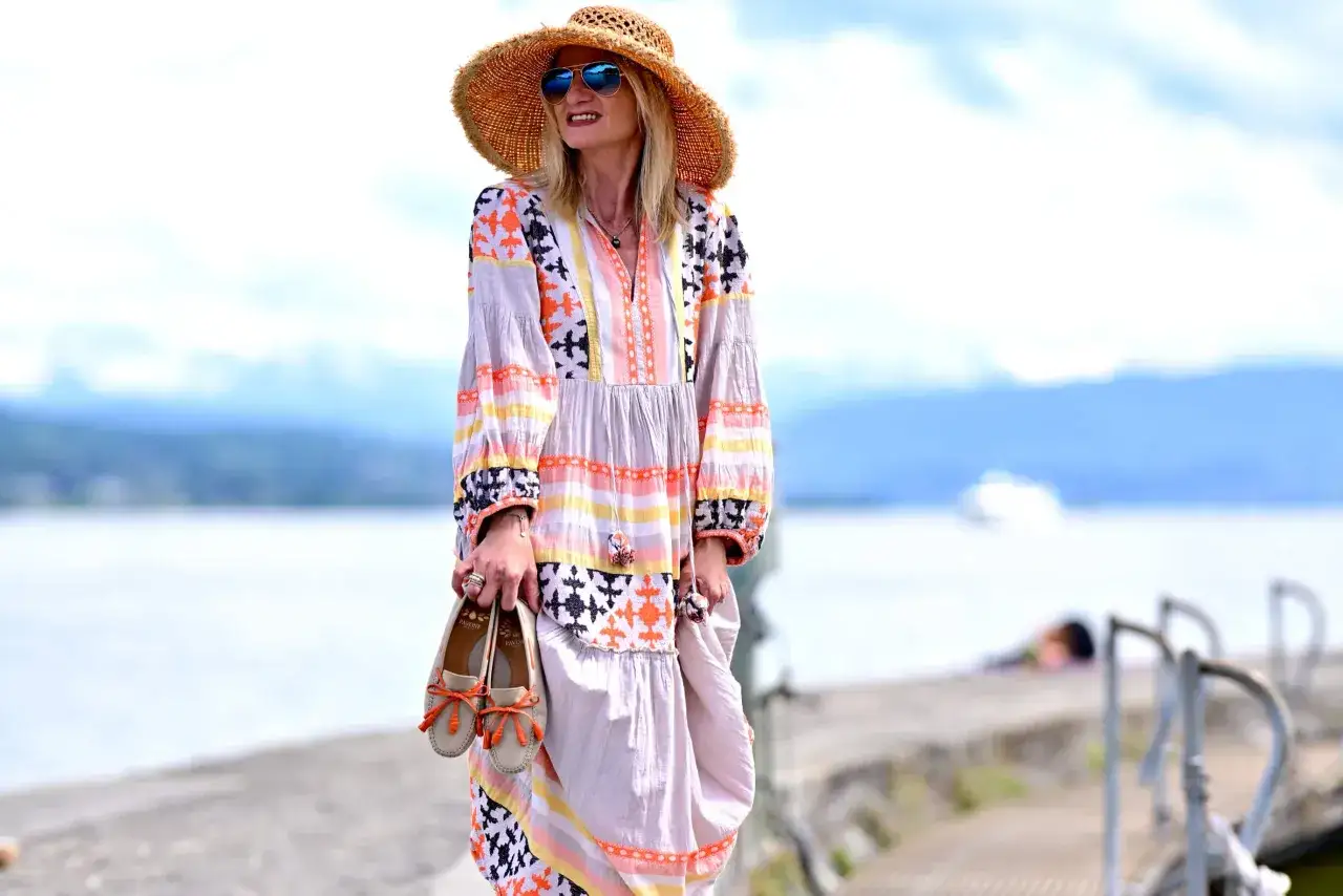 Kobieta w sukience boho i słomkowym kapeluszu trzyma beżowe mokasyny z pomarańczowymi kokardkami. Idealne buty do sukienki boho na letni spacer.