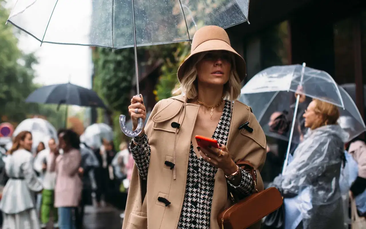 A woman in a stylish camel coat and bucket hat holds a clear umbrella, showcasing aesthetic rainy day outfits amidst a crowd.