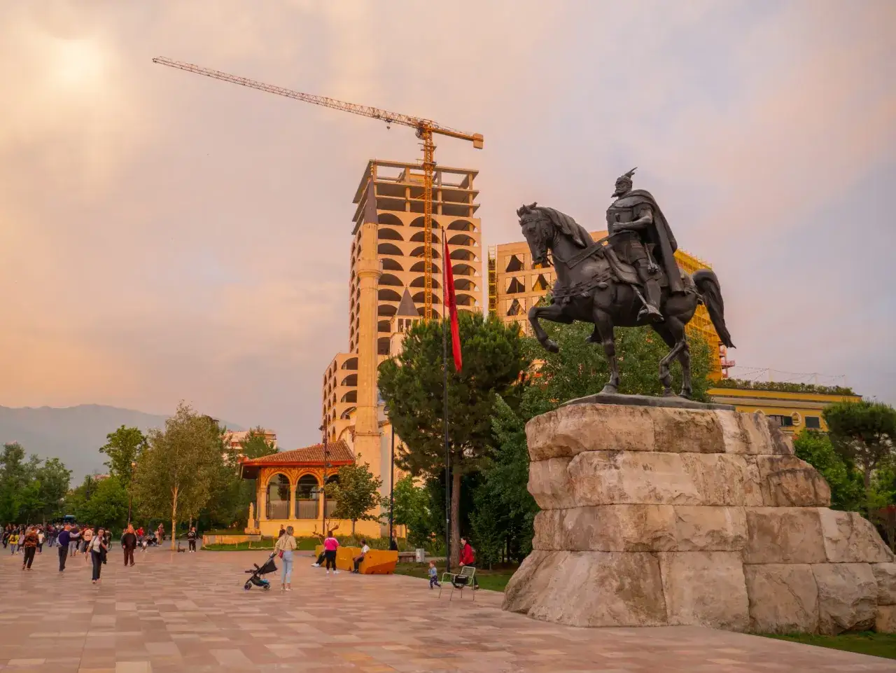 Pomnik Skanderbega na placu w Tiranie, w tle budynek w budowie i góry. Wiele osób spaceruje, odkrywając tirana atrakcje.