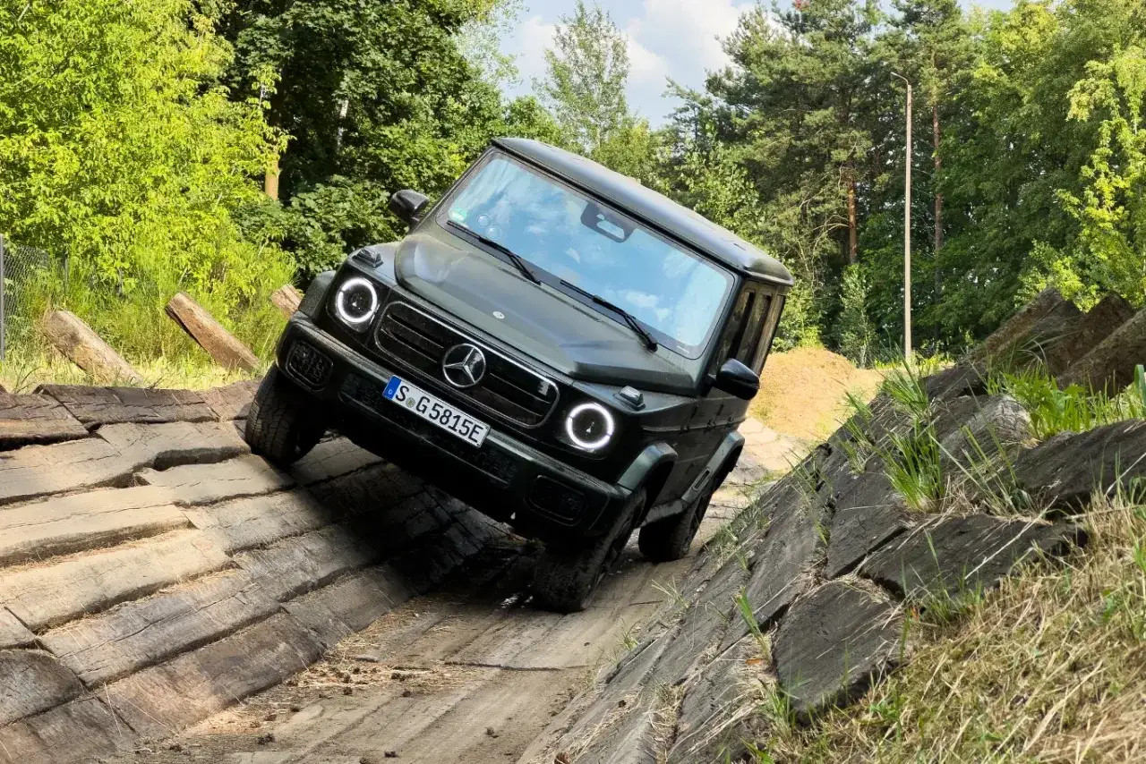 Ciemnozielony Mercedes Klasy G pokonuje trudny teren, wspinając się po drewnianej konstrukcji.
