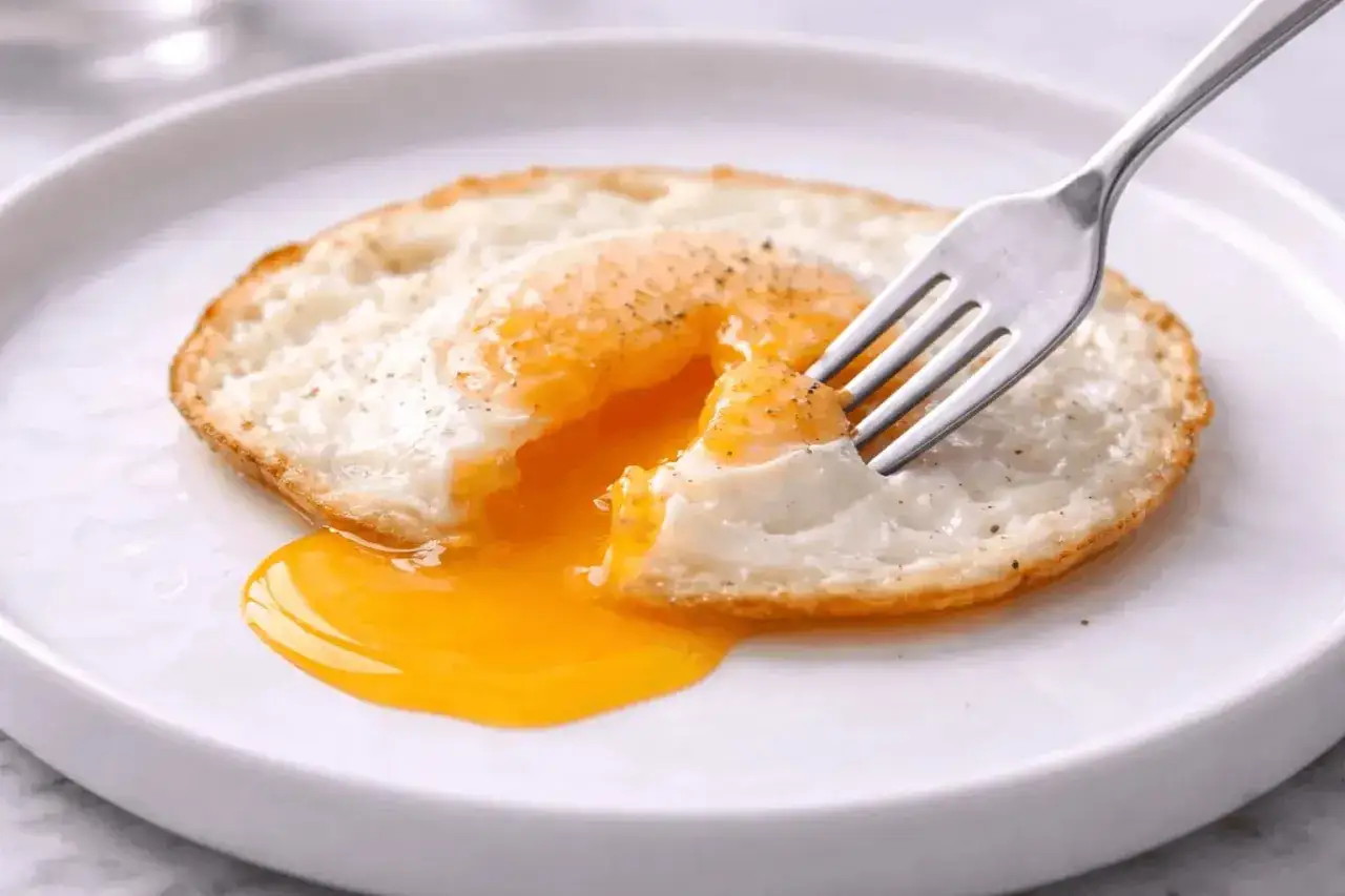 Un &oelig;uf miroir, le jaune coulant comme de l'or liquide, est perc&eacute; par une fourchette sur une assiette blanche.
