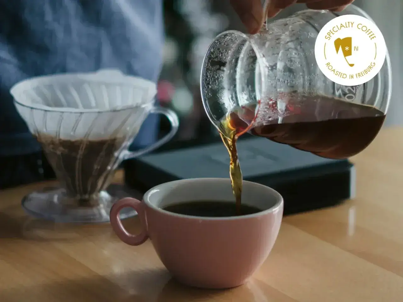 Frisch aufgebr&uuml;hter Kaffee flie&szlig;t aus einer Glaskaraffe in eine rosa Tasse. Der **blooming effekt** des Kaffees ist sichtbar.