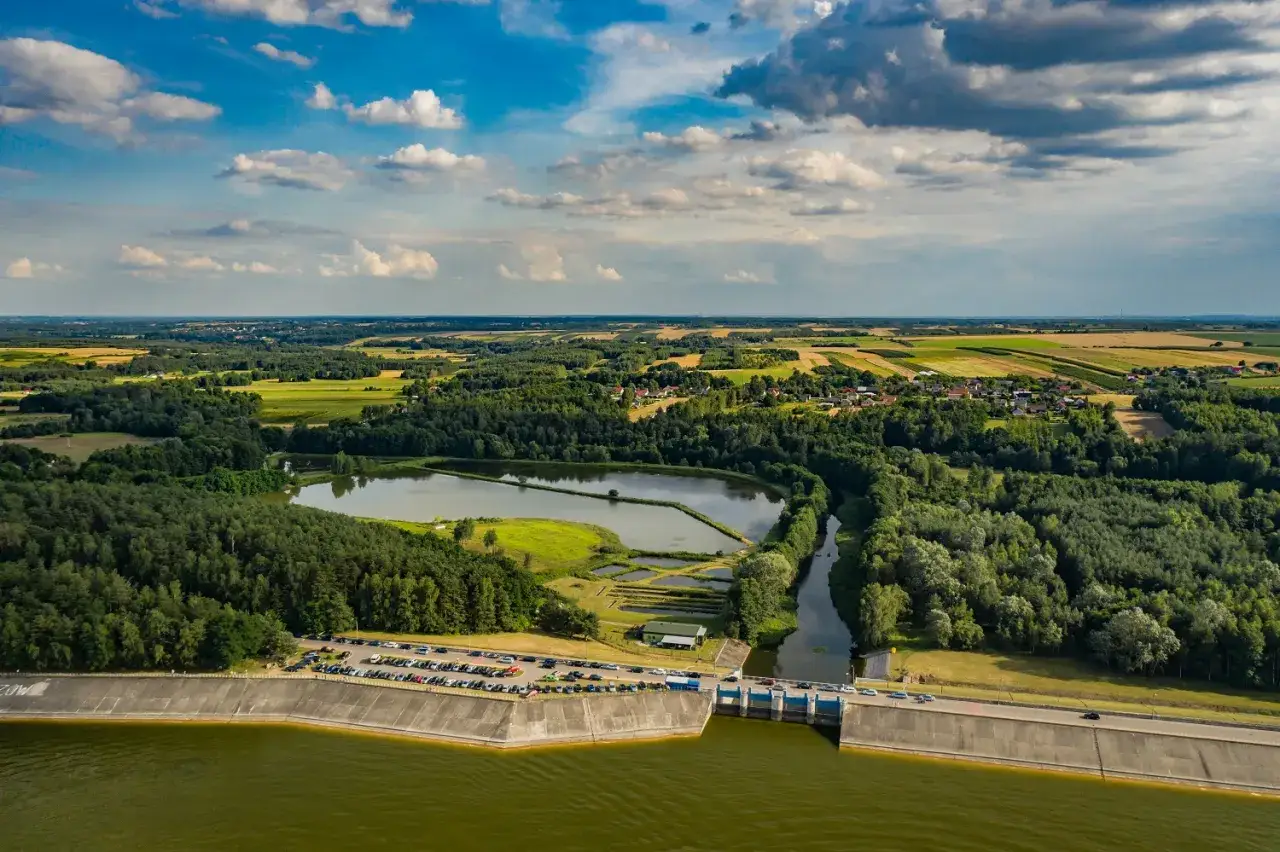 Widok z lotu ptaka na chańcza jezioro, otoczone lasami i polami. Na pierwszym planie betonowa zapora, za nią spokojna tafla wody.