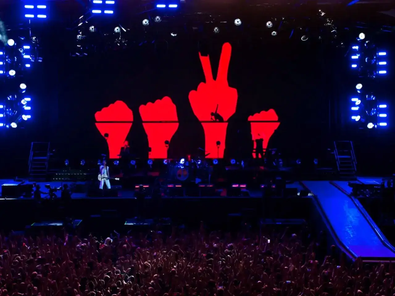 Koncert Depeche Mode. Na scenie wielkie czerwone dłonie, jedna z nich pokazuje znak pokoju. Tłum szaleje, słuchając ich depeche mode utwory.