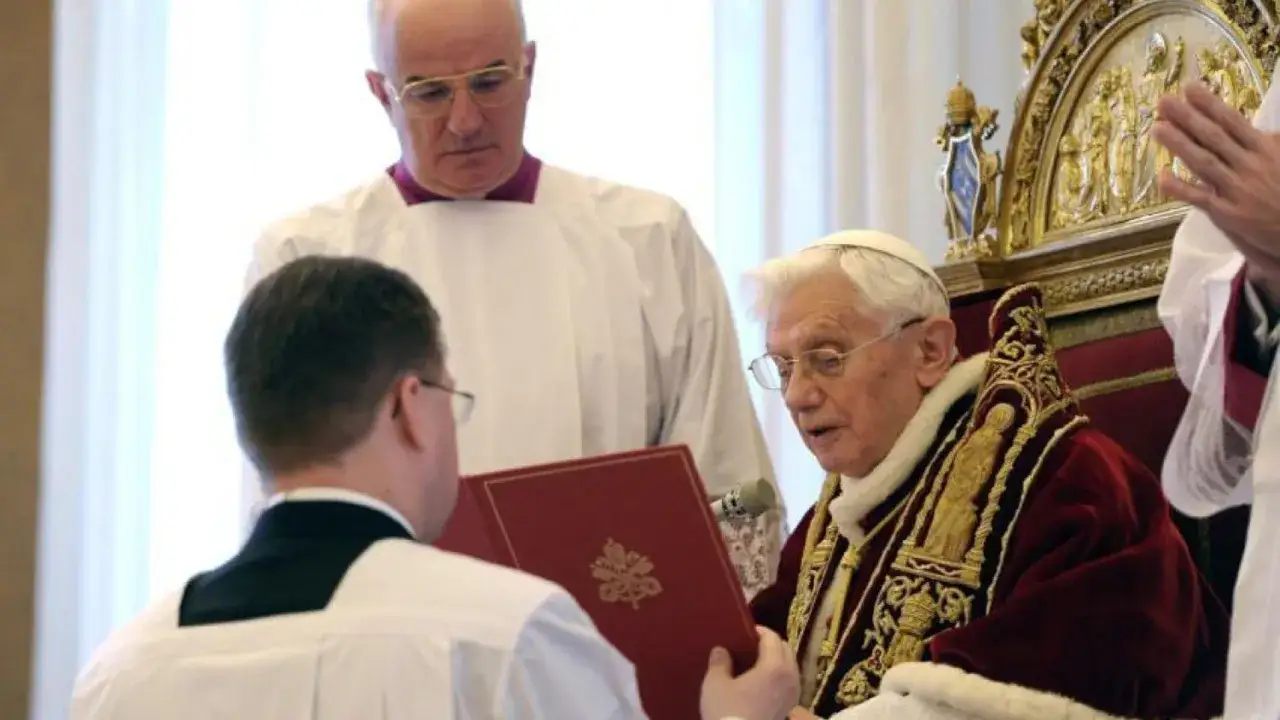 Papst Benedikt XVI. sitzt auf seinem Thron und erh&auml;lt ein Buch von einem Diakon. Ein weiterer Geistlicher steht hinter ihm.