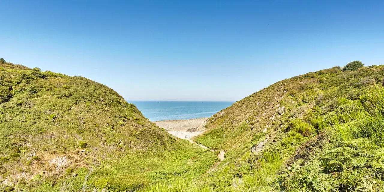 Vue sur la mer à travers la vallée du Lude, bordée de collines verdoyantes et d'une plage de sable.