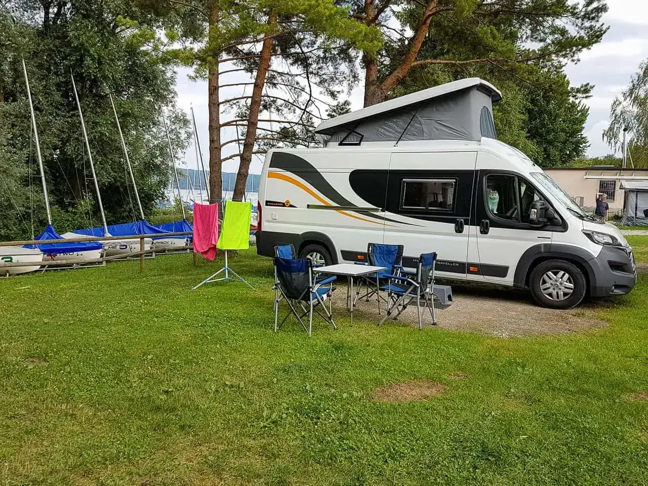 Kamper gotowy na wakacje, z rozłożonym stolikiem i krzesłami. W tle żaglówki i suszące się ręczniki – idealne gadżety do kampera na wypoczynek nad wodą.