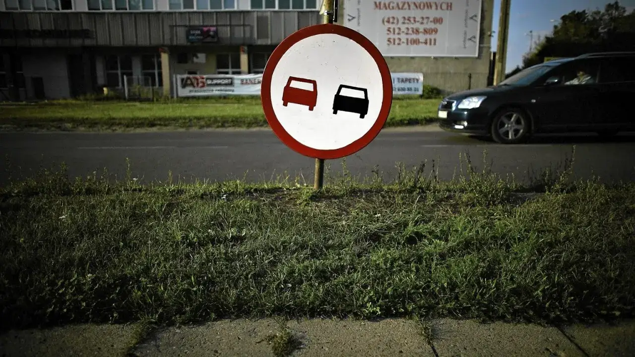 Znak zakazu wyprzedzania. Dwa samochody, jeden czerwony, drugi czarny, symbolizują zakaz **wymijania** i **omijania**.