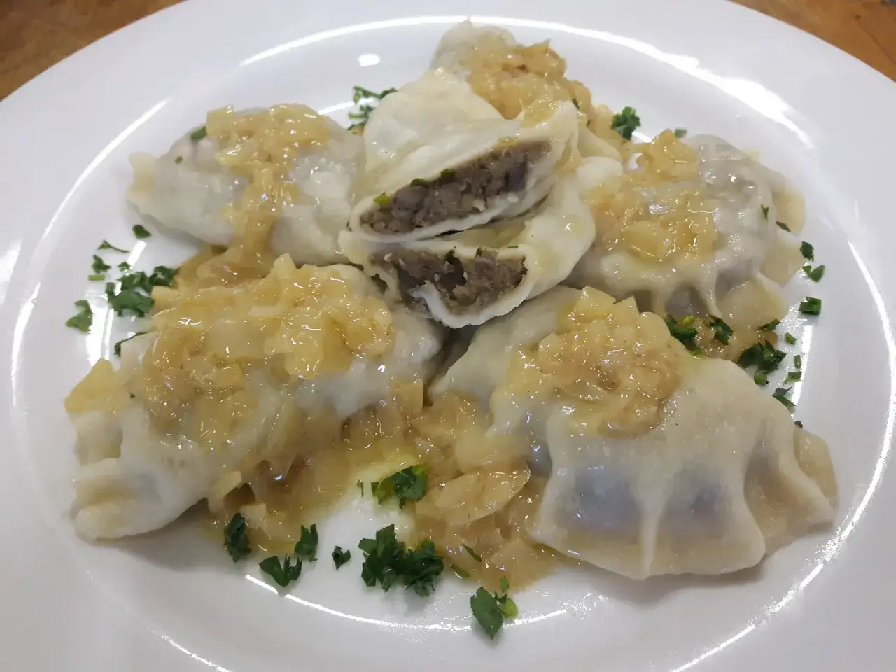 Pyszne pierogi z soczewicą, polane skarmelizowaną cebulką i posypane natką pietruszki, prezentują się apetycznie na białym talerzu.