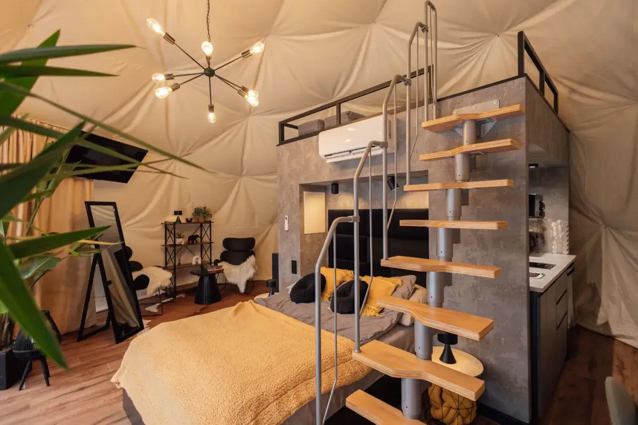 Nowoczesny glamping w Nałęczowie: przestronne wnętrze kopuły z łóżkiem, schodami na antresolę i aneksem kuchennym.