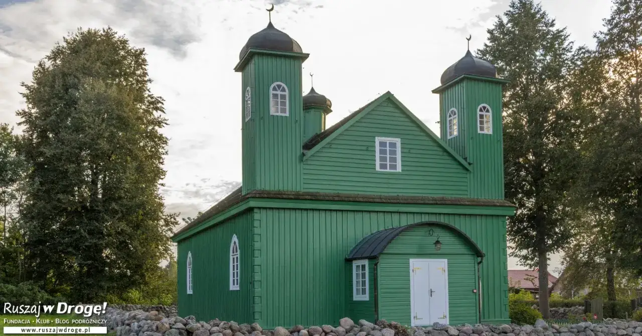 Zielony, drewniany meczet z minaretami i księżycami na szczytach. Otoczony kamieniami i drzewami.