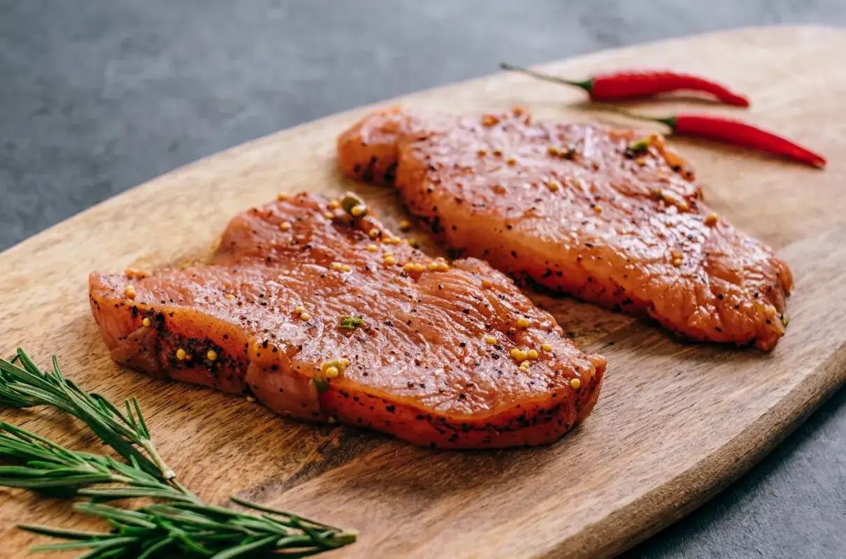 Dwa kawałki indyka w marynacie z gorczycą i pieprzem, obok gałązka rozmarynu i dwa czerwone chili.