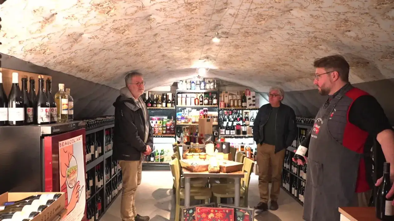 Trois hommes dans une cave à vin, entourés de bouteilles. L'un d'eux tient une bouteille, expliquant peut-être le terroir.