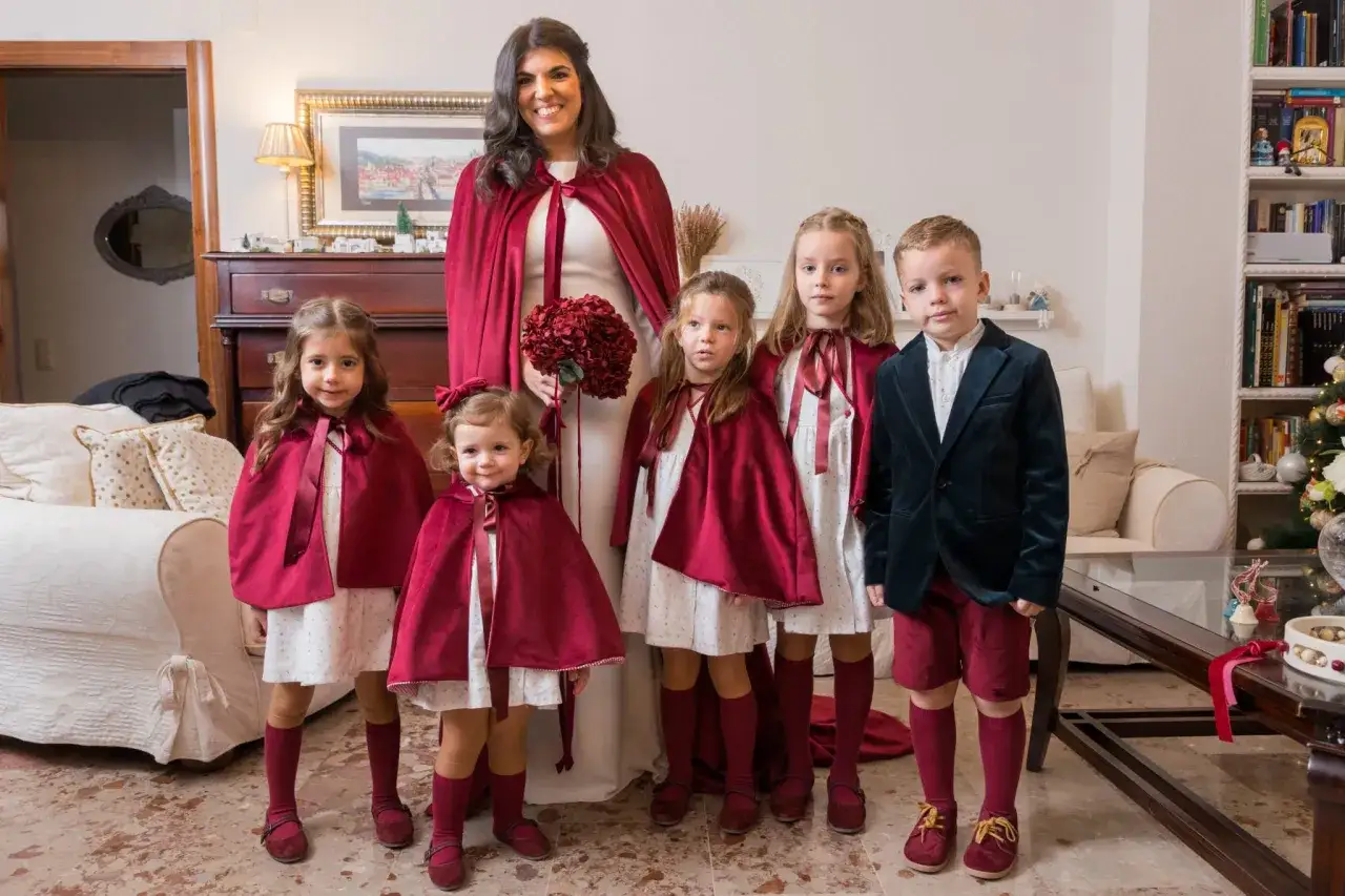Novia sonriente con ramo y cinco pajes boda con capas burdeos.