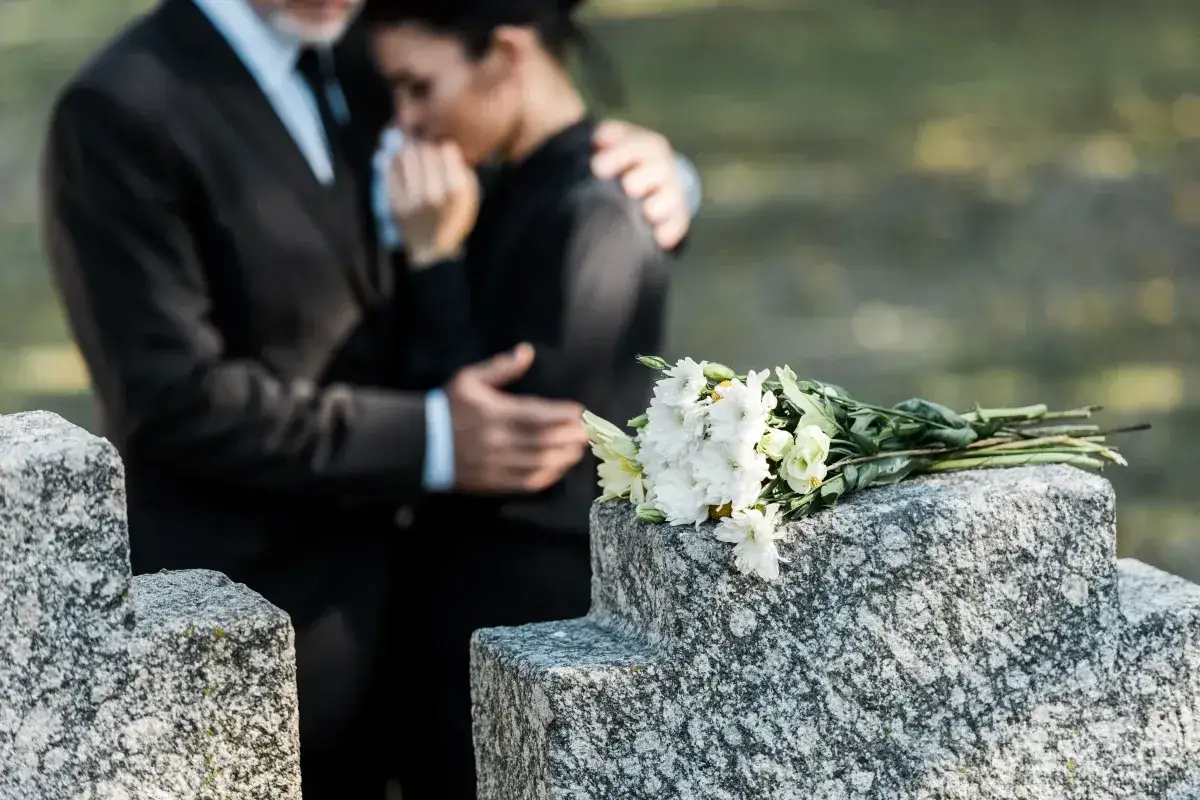 Para, obejmująca pogrążoną w żalu kobietę, przy grobie z kwiatami. Trudny czas, jak urlop macierzyński po poronieniu, wymaga wsparcia.