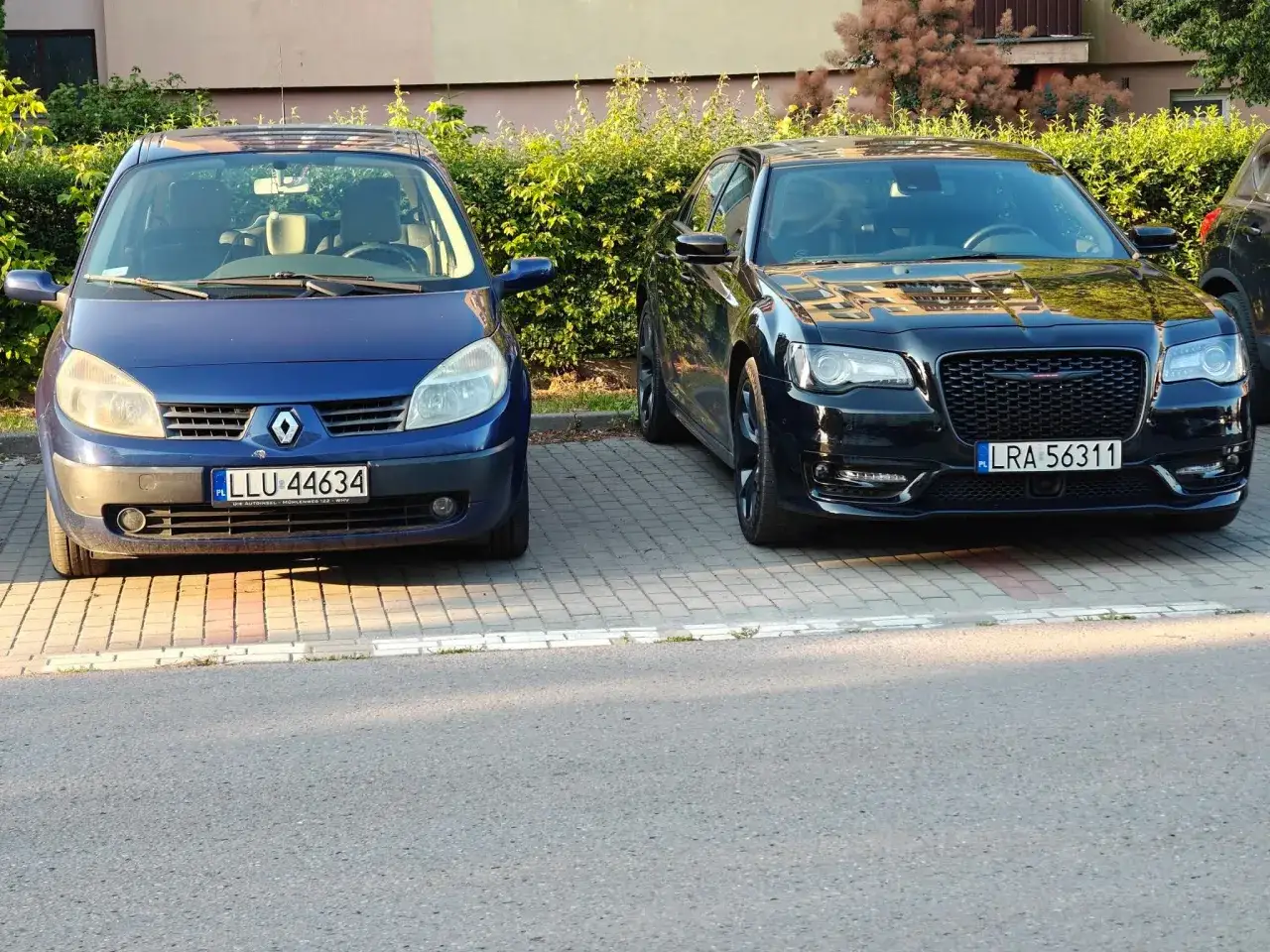 Dwa samochody: niebieski Renault Scenic z tablicą LLU 44634 i czarny Chrysler z LRA 56311, zaparkowane obok siebie.