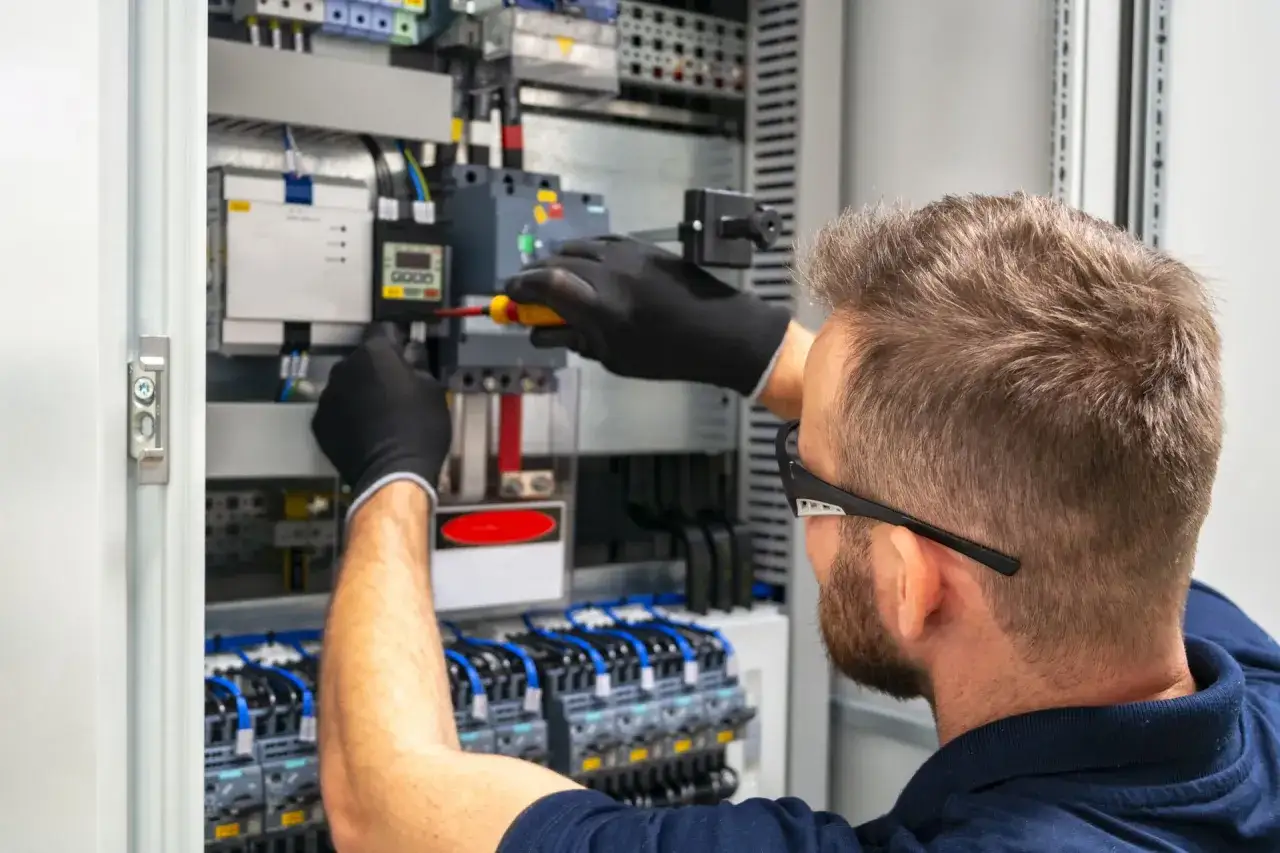 Elektryk w rękawiczkach pracuje przy panelu sterowania. Zastanawiasz się, ile kosztuje godzina pracy elektryka?