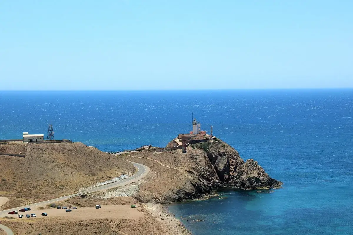 La cala del Corralete, con su faro en la cima rocosa, se baña en el azul intenso del mar Mediterráneo. Una carretera serpentea hacia la costa.