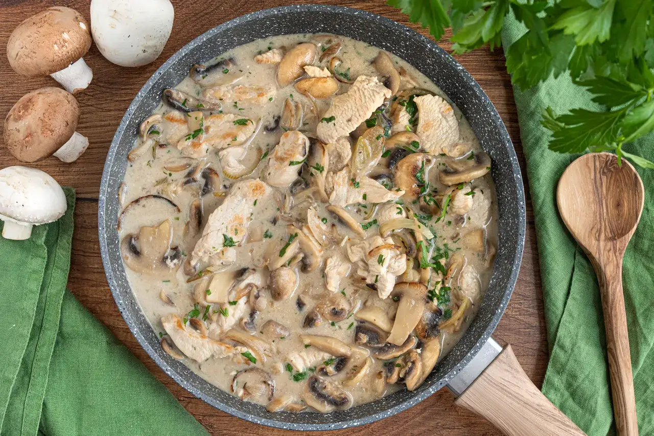 Herzhaftes Puten Geschnetzeltes mit Pilzen in cremiger Soße, garniert mit Petersilie. Daneben frische Champignons und ein Holzlöffel.