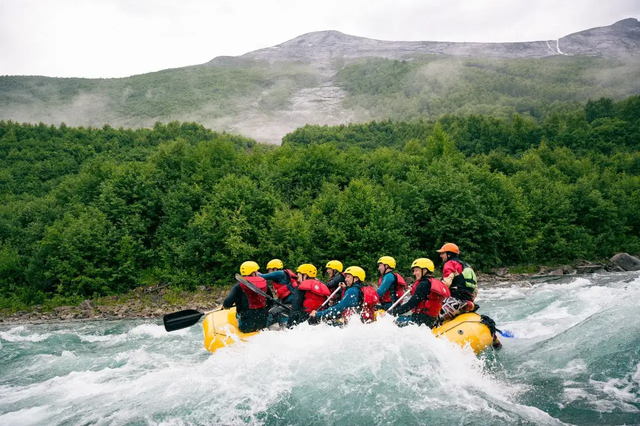 En gul flotte med åtta personer paddlar genom forsen. Sjoa rafting i en dramatisk bergsmiljö med grönskande skog.