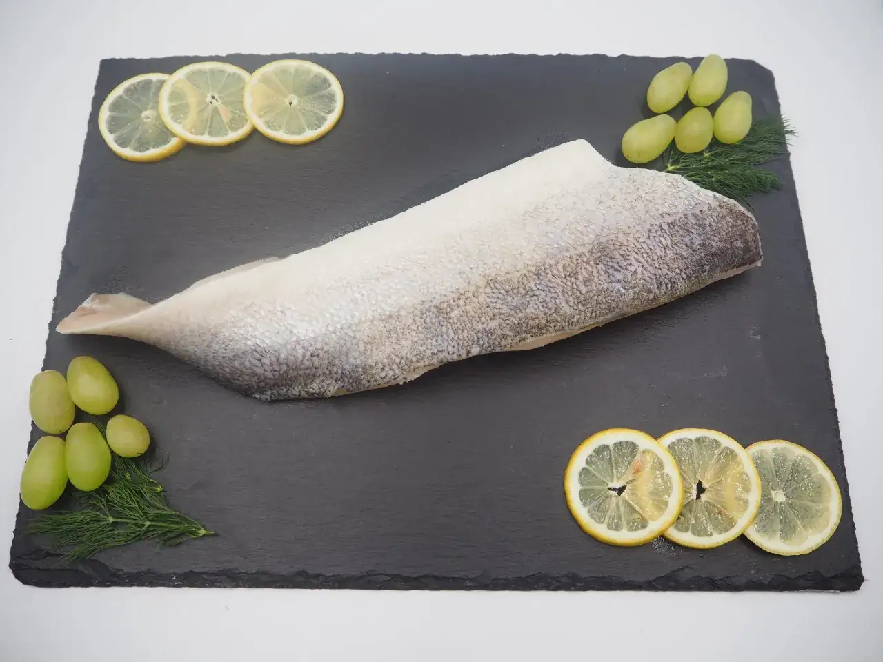 Filet sandacza, jaka to ryba, prezentowany z plasterkami cytryny, winogronami i koperkiem na czarnej desce.