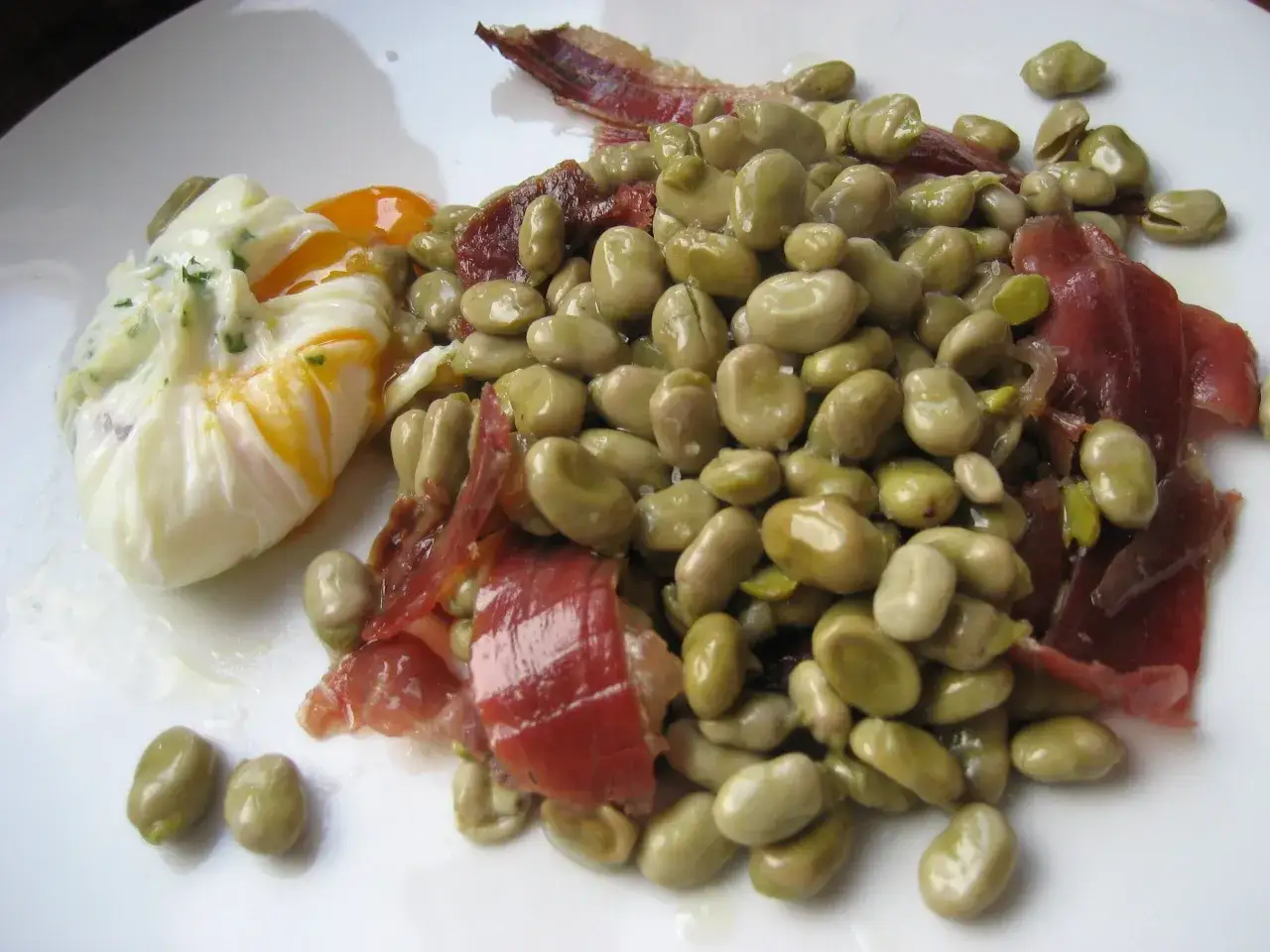 Un plato con habas tiernas, trozos de jamón serrano y un huevo escalfado con la yema a medio cuajar.