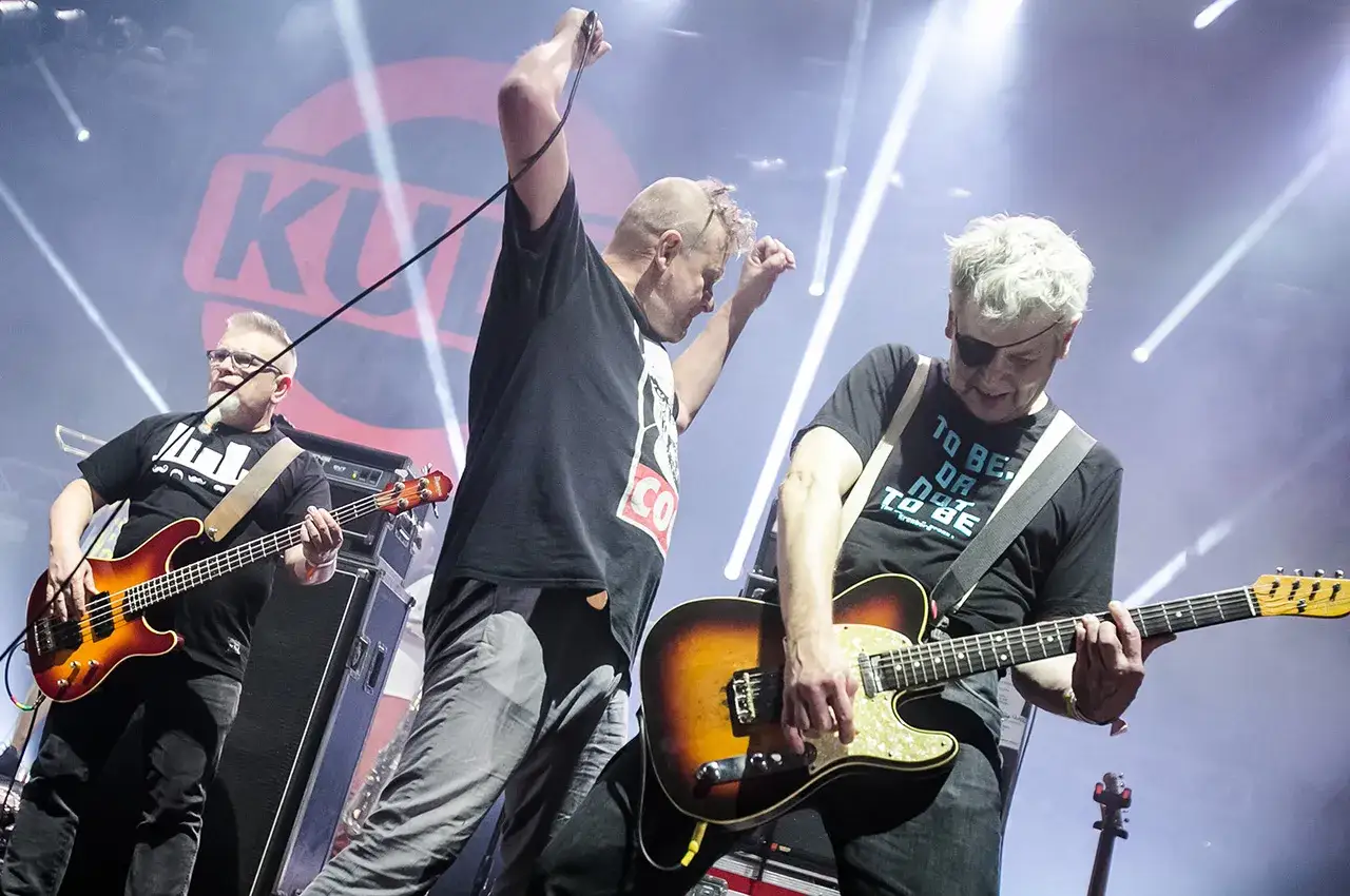 Zespół Kult na koncercie, grający swoje kultowe utwory. Wokalista z mikrofonem, gitarzysta z gitarą i basista.