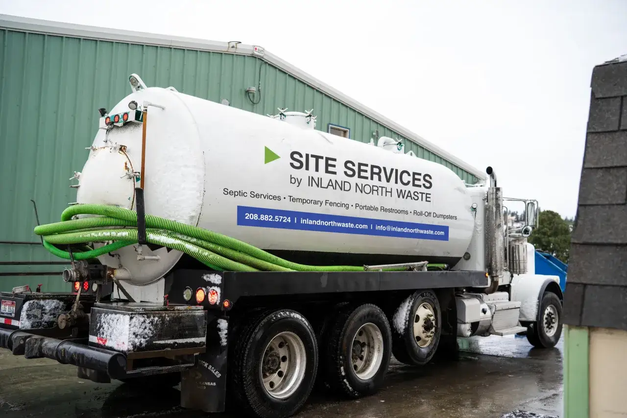 A white septic system maintenance truck with green hoses is parked outside a building.