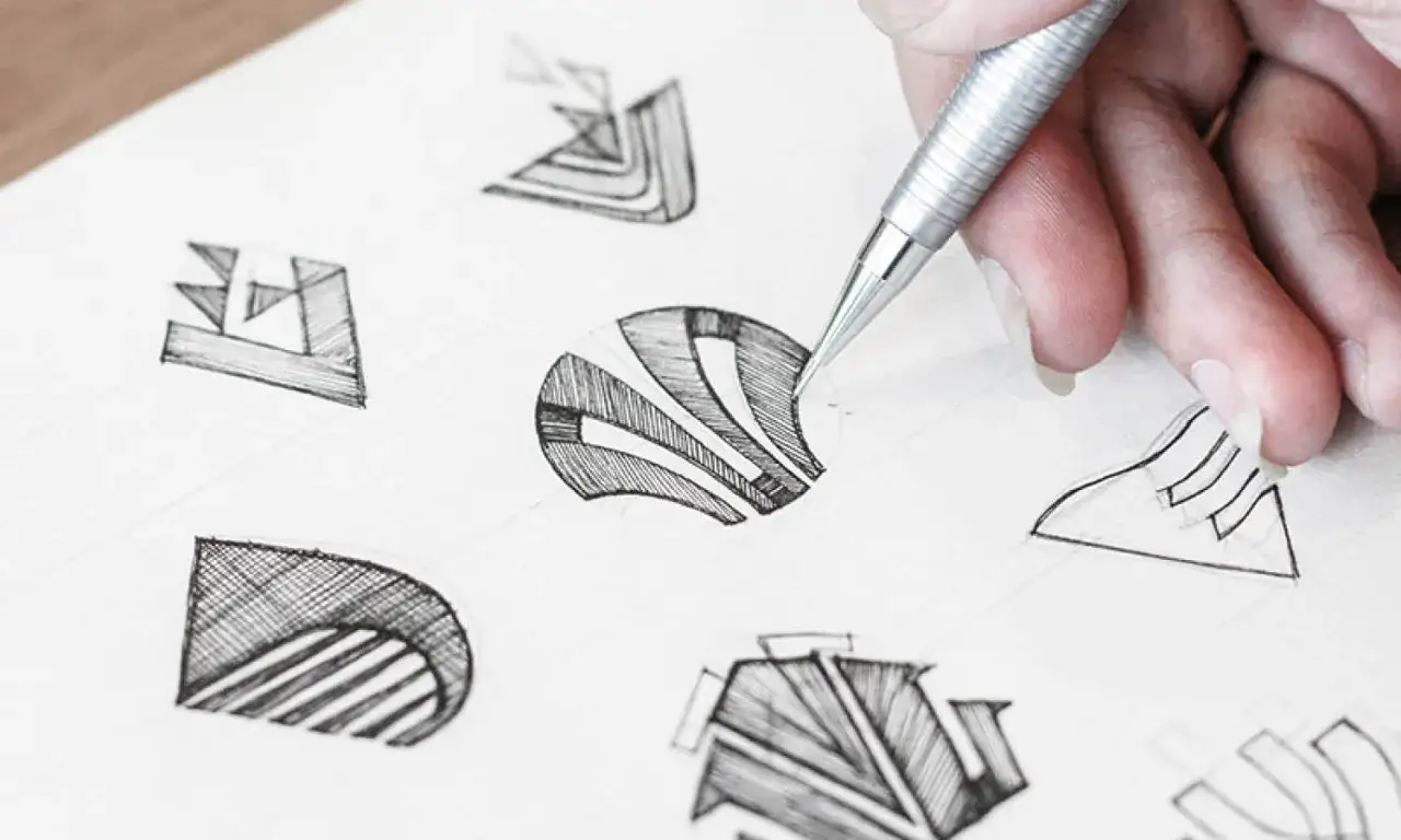 Main d'un designer esquissant des id&eacute;es pour comment dessiner un logo, avec plusieurs croquis g&eacute;om&eacute;triques au crayon sur papier.