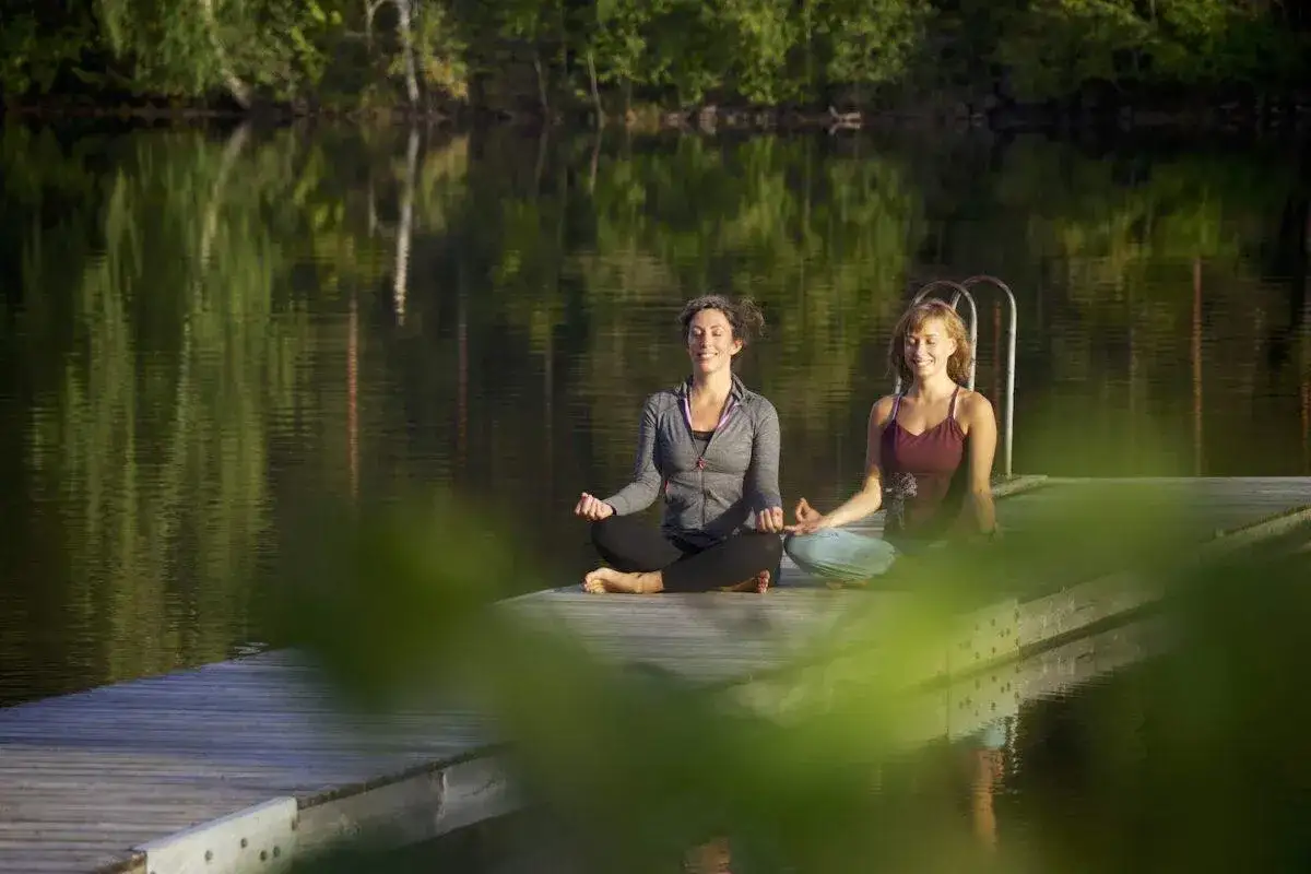 Två kvinnor mediterar på en brygga vid en spegelblank sjö. En perfekt yoga helg i naturen.