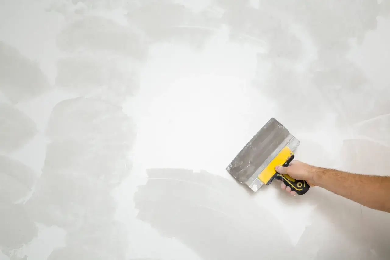 Mano con espátula amarilla y gris alisando una pared con gotelé, mostrando cómo quitar el gotelé sin esfuerzo.