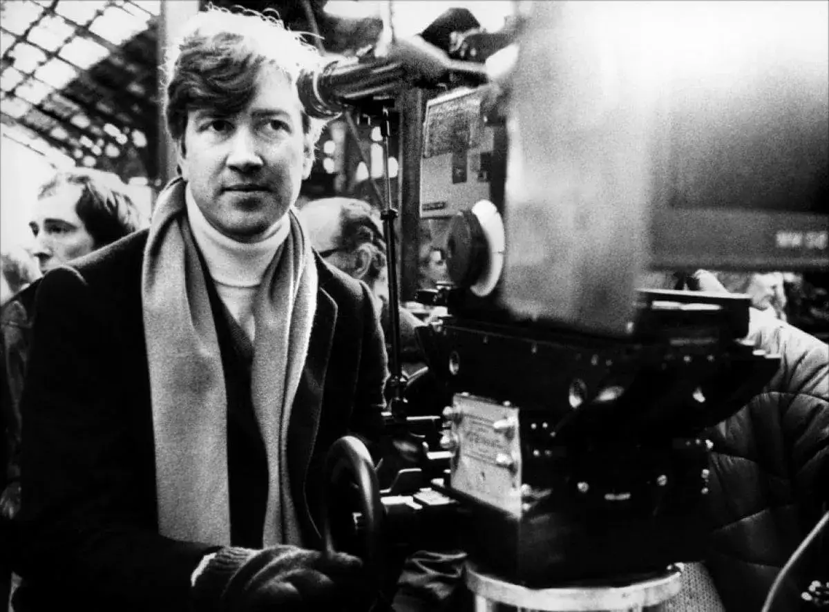 Młody David Lynch z charakterystycznym szalikiem i golfem, stoi obok kamery filmowej.