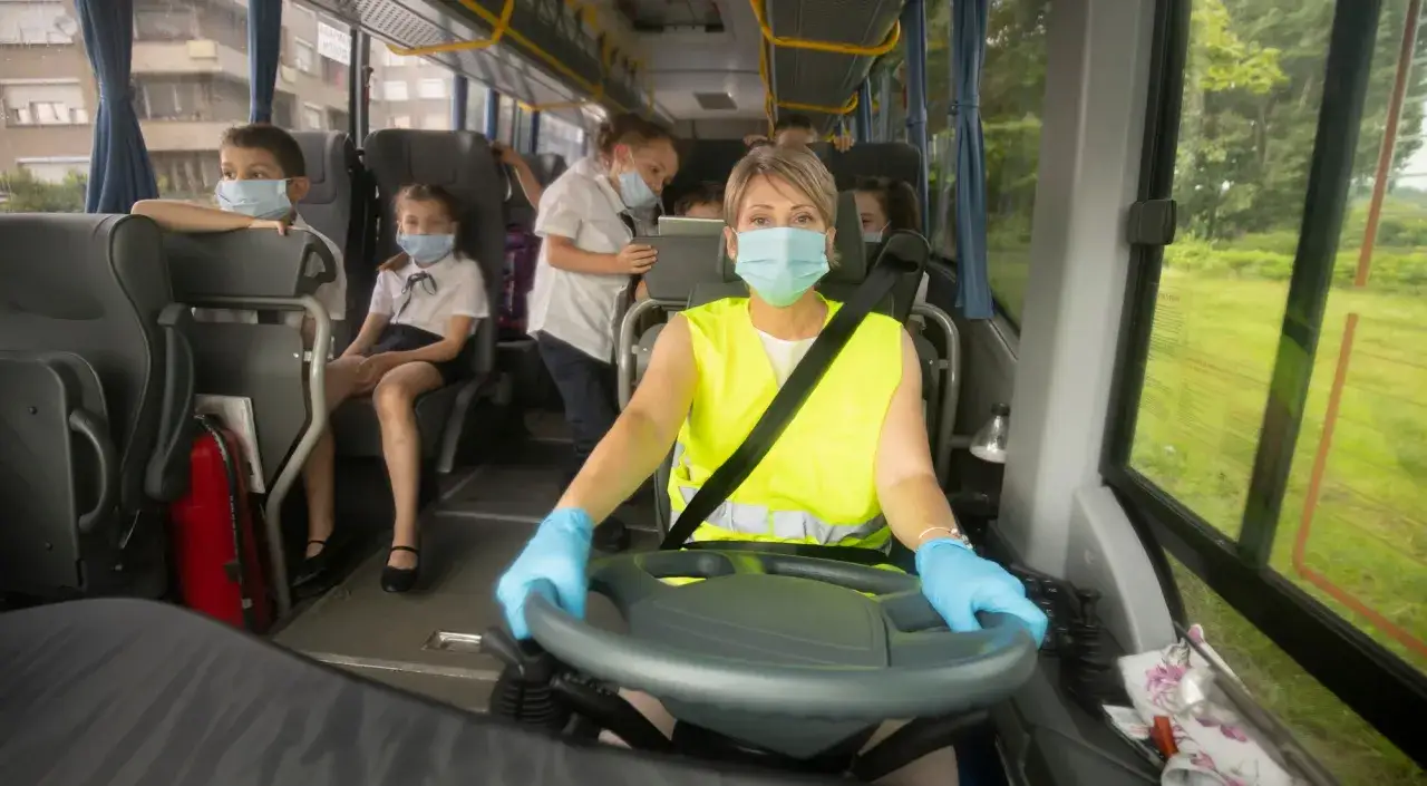 Conductora con mascarilla y guantes, supervisa a ni&ntilde;os con mascarillas en el transporte escolar, cumpliendo la normativa.