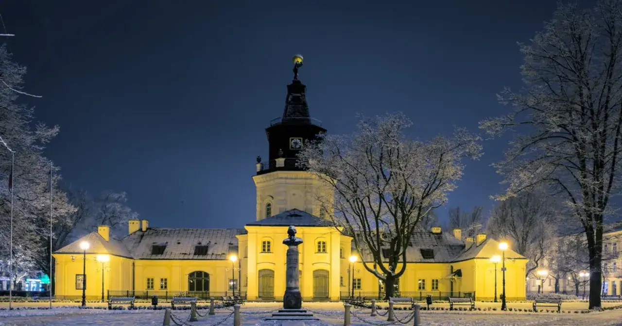 Zimowa noc w Siedlcach. Oświetlone zabytki, w tym pałac i wieża z zegarem, otoczone ośnieżonymi drzewami.