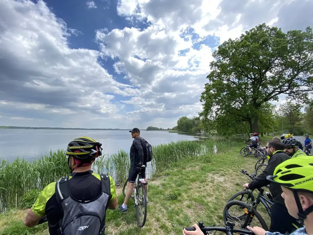 Grupa rowerzyst&oacute;w odpoczywa nad jeziorem, podziwiając malownicze **stawy milickie trasy rowerowe**.
