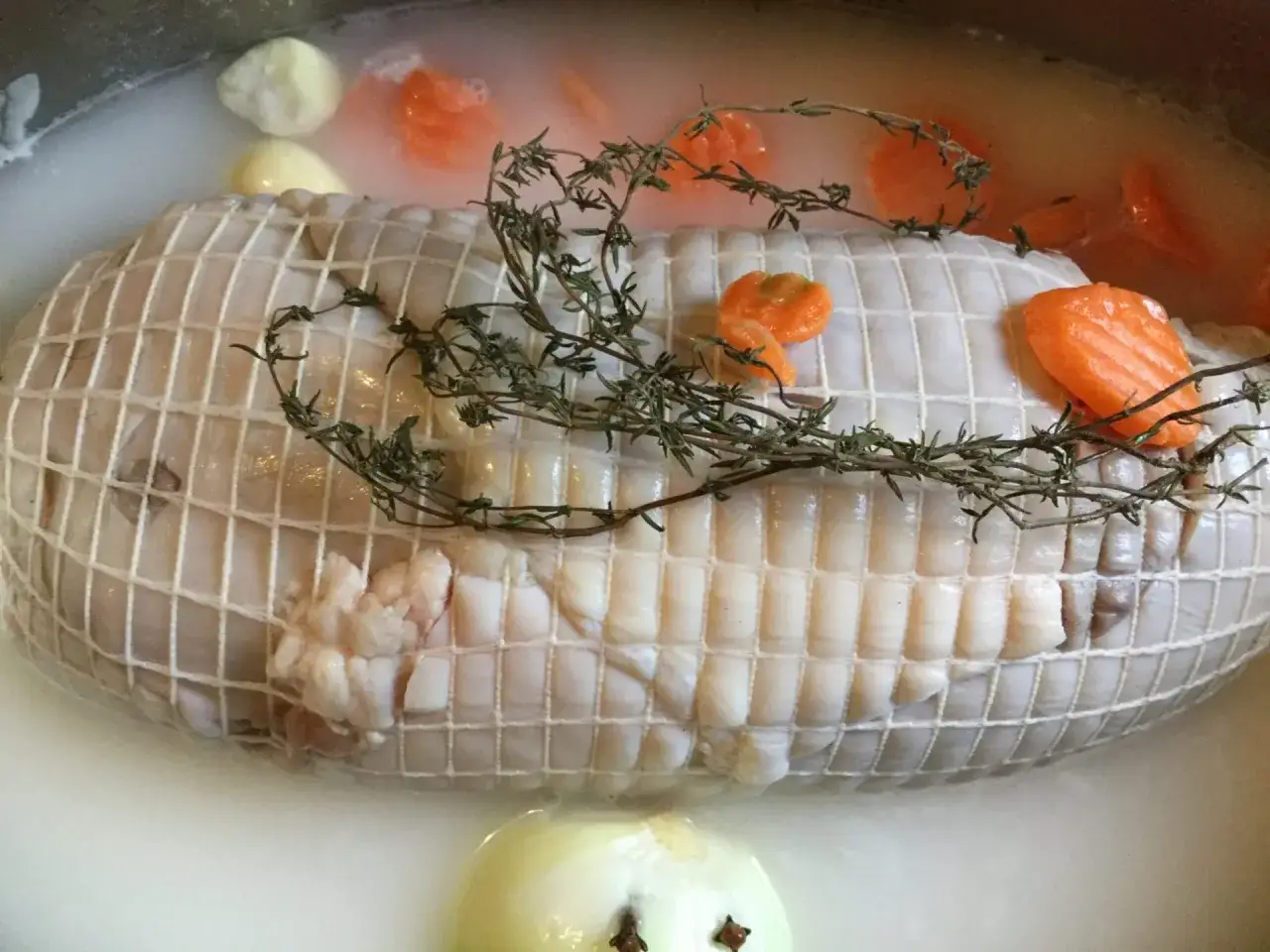Préparation de la cuisson tête de veau cocotte. Le veau est ficelé, entouré de carottes et de thym dans un bouillon.