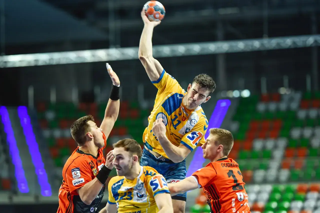 Gracz w żółtej koszulce rzuca piłkę do bramki, broniony przez zawodników w pomarańczowych strojach. Emocjonujący moment z meczu final four handball.