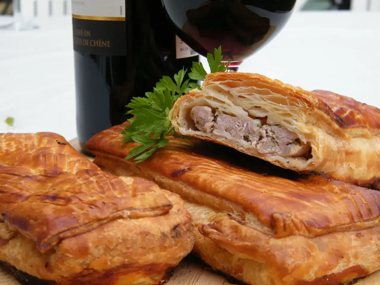 Pâté en croûte, une spécialité lorraine savoureuse, avec une garniture de viande visible. Une bouteille de vin rouge et du persil complètent la scène.