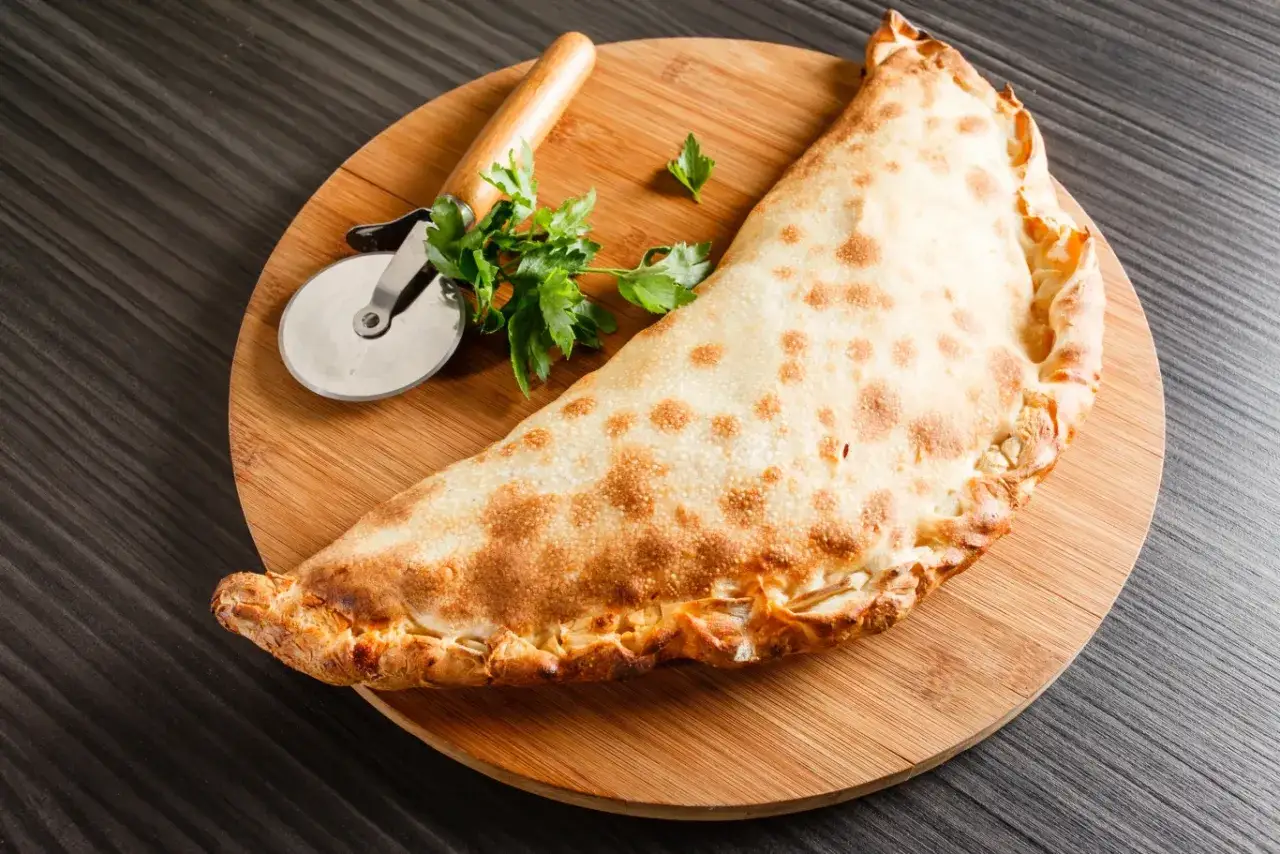 Złocisty calzone, czyli włoska pierożkowa pizza, leży na desce z krążkiem do pizzy i natką pietruszki.