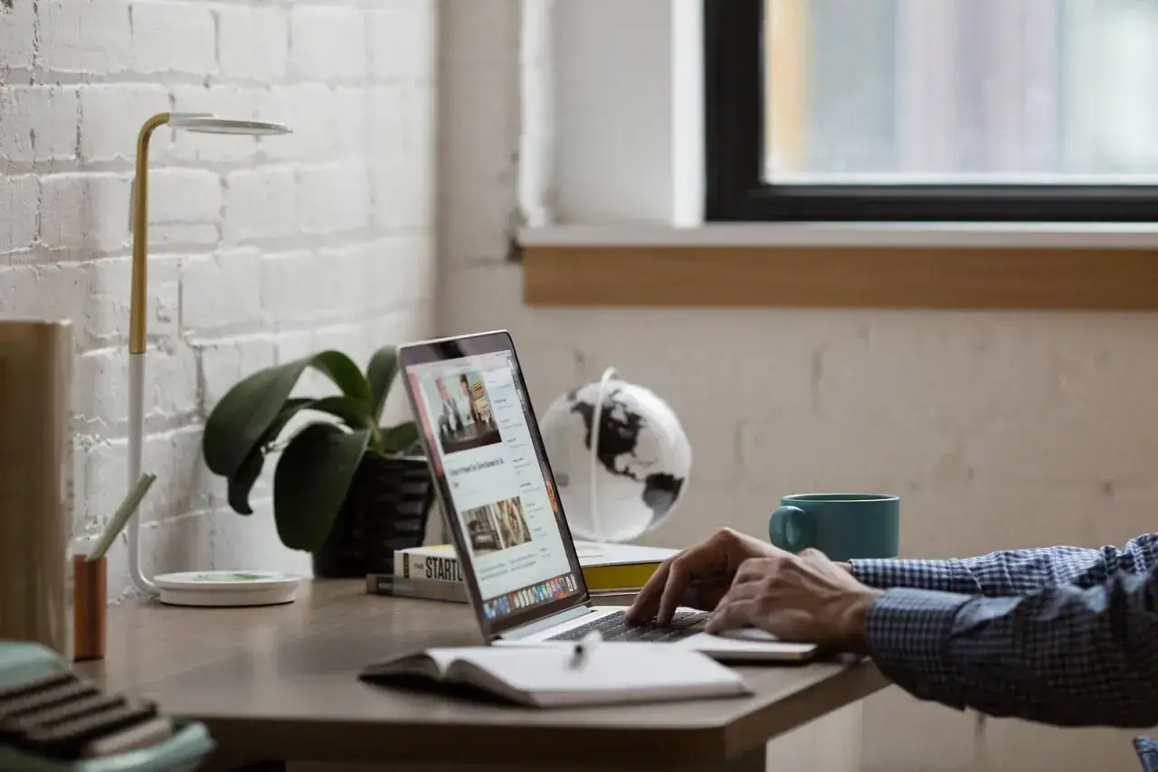 Person tippt auf Laptop, daneben stehen Lampe, Pflanze, Globus und Tasse.