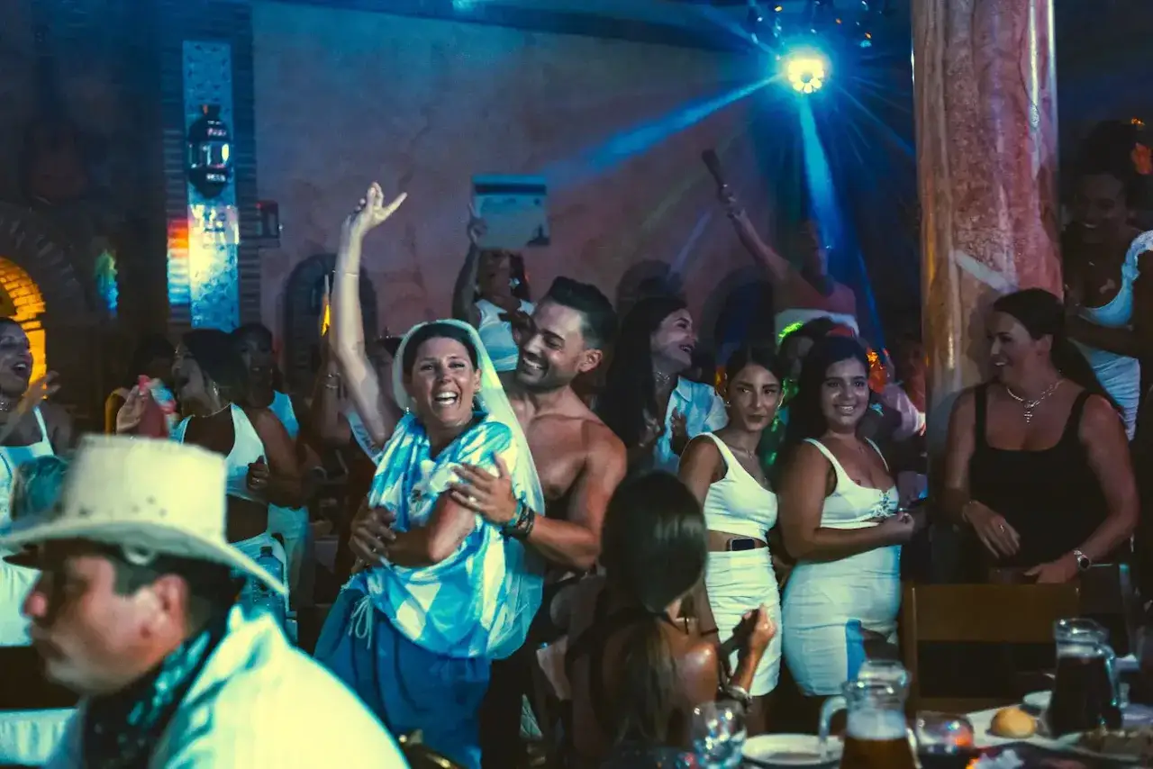 Gente celebrando y bailando en una fiesta, con luces de discoteca y un ambiente festivo. Parece una despedida de soltero con juegos y mucha alegría.