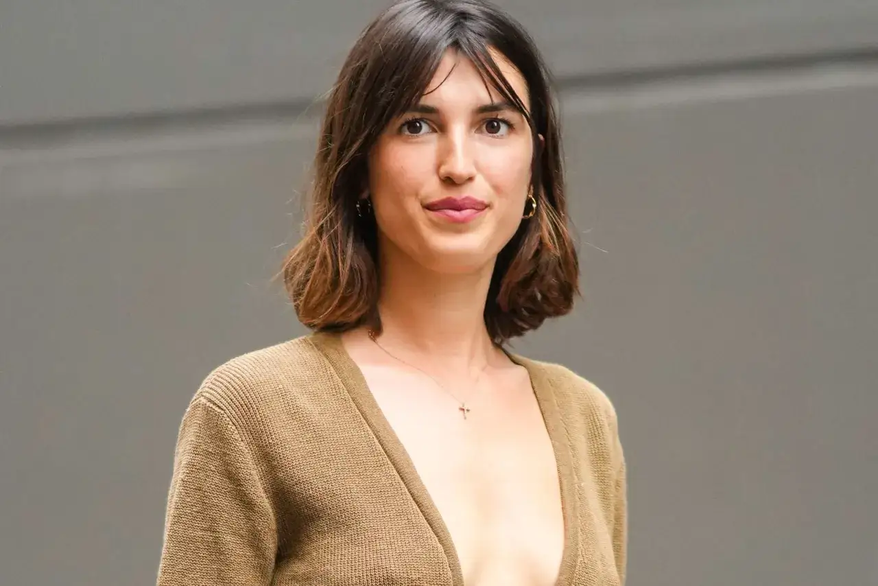 Mujer con cabello castaño ondulado, en transición de pelo corto a largo, luce un suéter marrón y un collar con cruz.