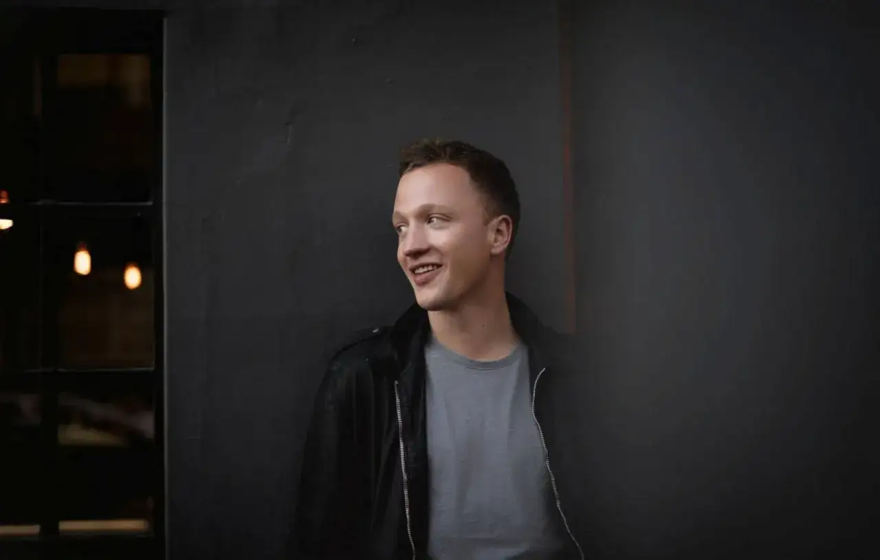Joris, lächelnd, lehnt an einer dunklen Wand.