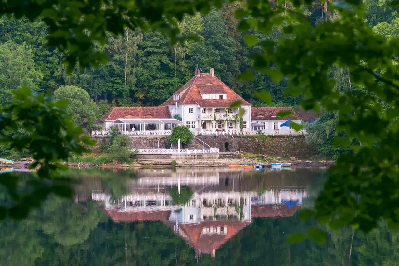 Dom z czerwonym dachem nad jeziorem, odbijający się w wodzie, otoczony zielenią.