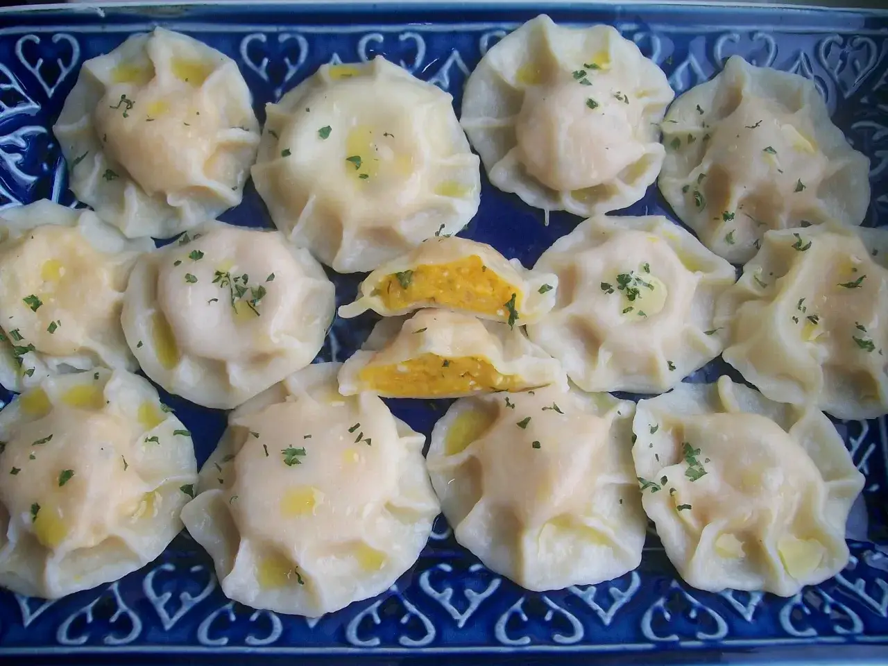 Pyszne pierogi z dynią, polane masłem i posypane szczypiorkiem, ułożone na niebieskiej paterze.