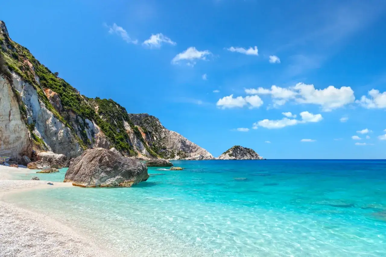 Słoneczna plaża z białym żwirem i turkusową wodą. W oddali skaliste zbocza porośnięte zielenią, charakterystyczne dla greckiej wyspy Kefalonia.