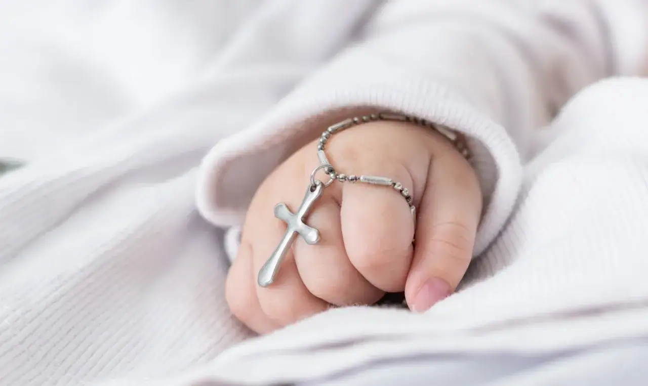 Dziecięca dłoń trzyma krzyżyk na łańcuszku, symbol tego, co jest potrzebne do chrztu.