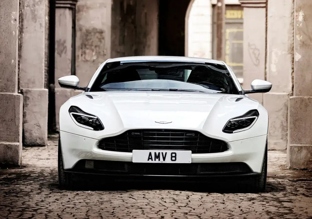 Biały Aston Martin DB11 z tablicą AMV 8 stoi na brukowanej uliczce, otoczony kamiennymi budynkami.