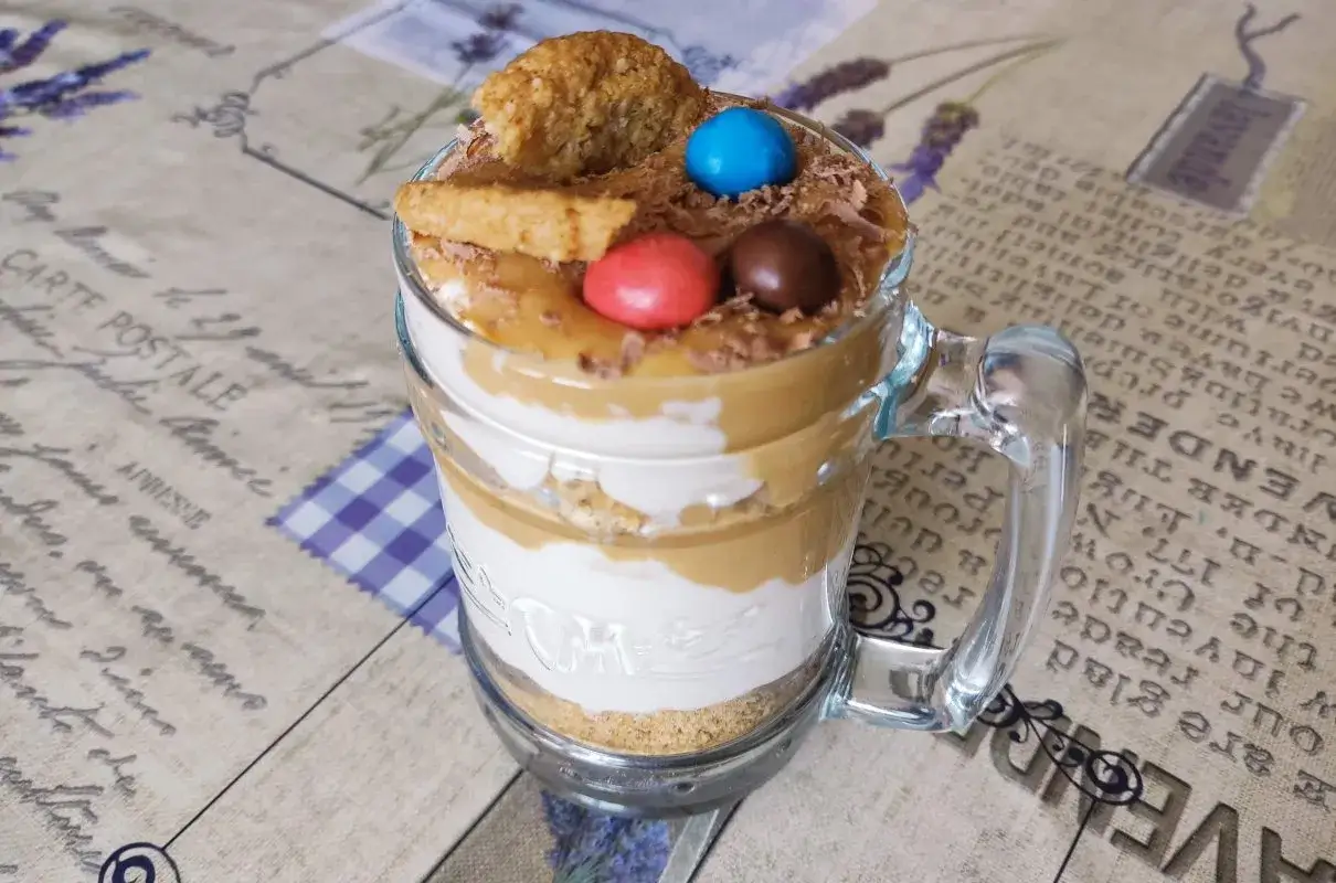 Warstwowy deser banoffee w szklance z polewą karmelową, bitą śmietaną, ciasteczkami i kolorowymi drażetkami.