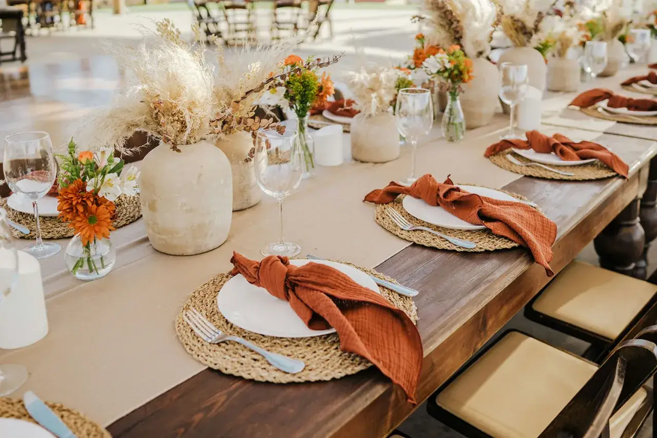 Un élégant dressage de table à la française avec des vases en céramique, des herbes séchées, des fleurs orangées et des serviettes en tissu rouille.